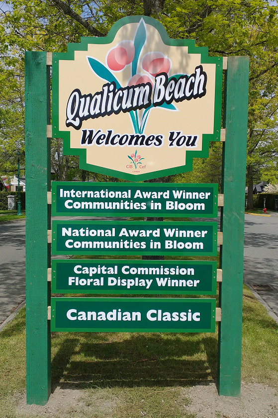 A green sign outside in a park that reads Qualicum Beach Welcomes You, International Award Winner Communities in Bloom, National Award Winner Communities in Bloom, Capital Commission Floral Display Winner
