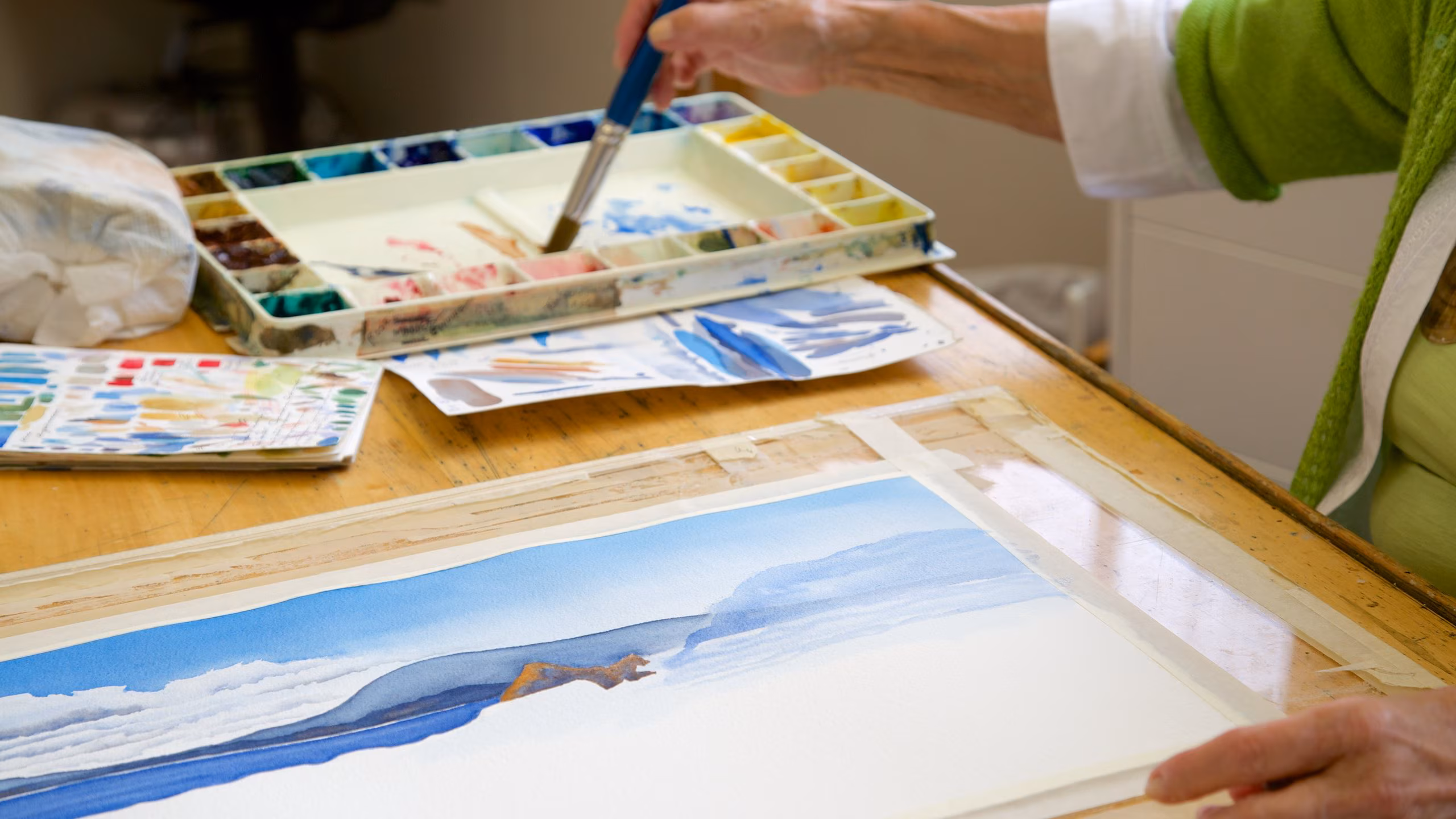A person is painting on a canvas, holding a paintbrush, with the artwork visible and in progress.