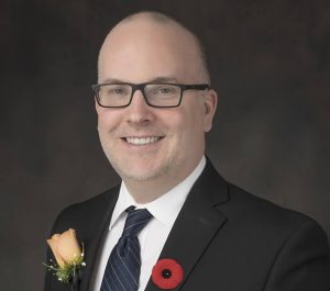 Councillor Harrison is pictured in front of a dark grey background. He wears dark-rimmed glasses, a dark suit, a plain white shirt, and a stiped navy tie. He wears a poppy pin on his left lapel and a light orange rose on his right lapel.