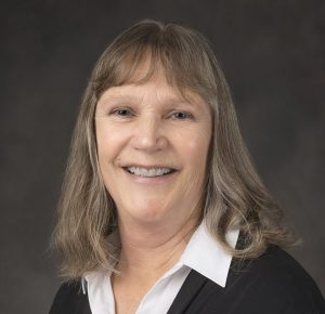 Councillor Vander Valk is pictured in front of a dark grey background. Her hair is down and comes to her shoulders. She wears a black cardigan and a white shirt.