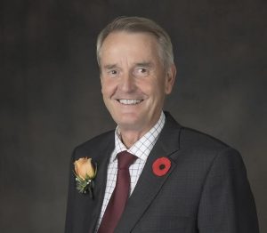 Mayor Teunis Westbroek against a dark grey background. Mayor Westbroek has short grey hair and is wearing a dark suit, checkered shirt, and burgundy tie. There is a red poppy pin on his left lapel, and a light orange rose on his right lapel.